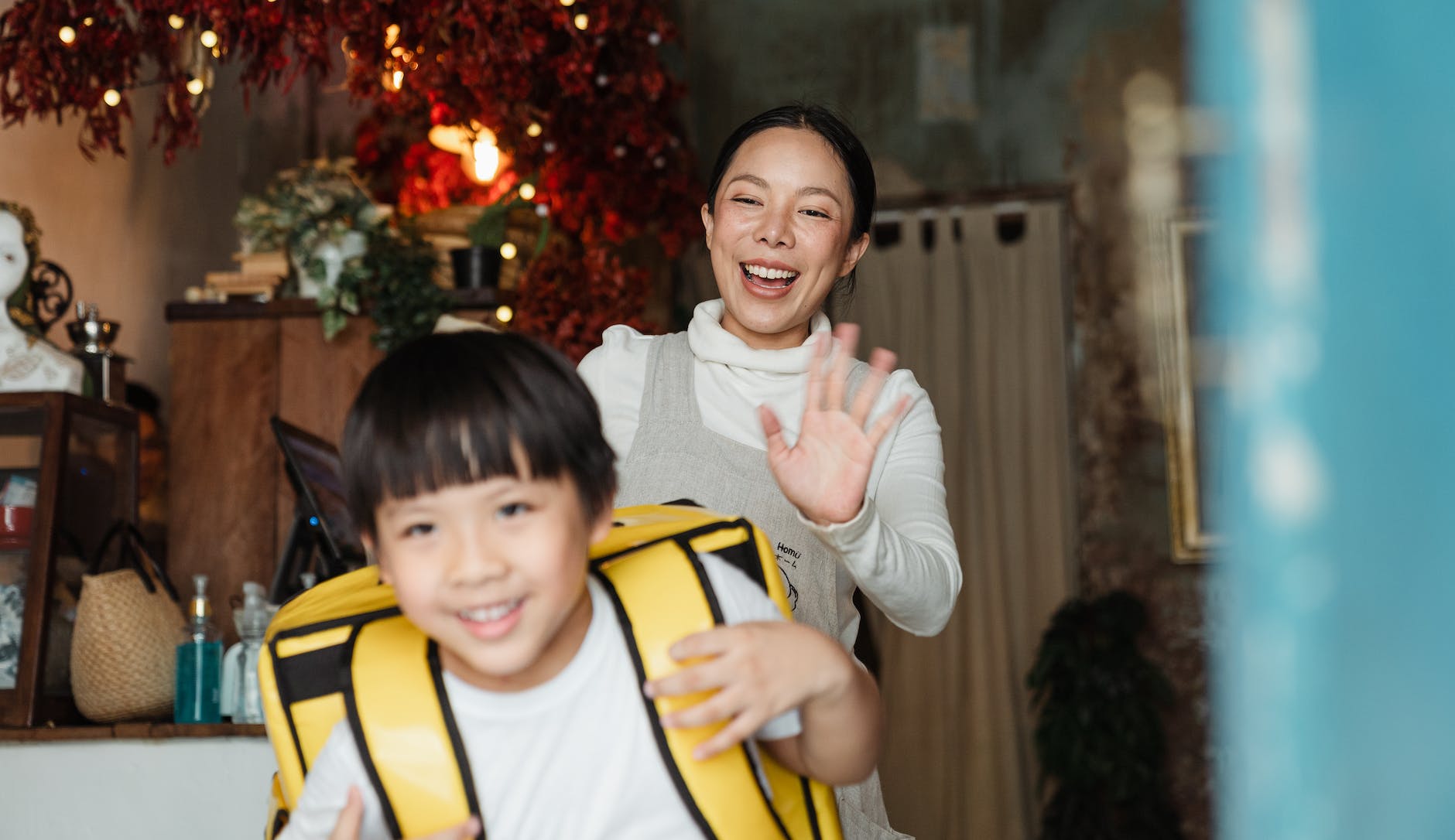 cheerful ethnic mother saying goodbye to son with food backpack